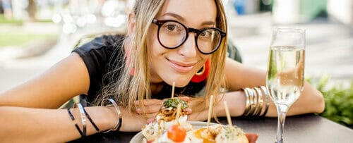 メガネをかけた女性が皿に料理とグラスワインを持ったテーブルに座り、スペイン風の食事を楽しんでいる。