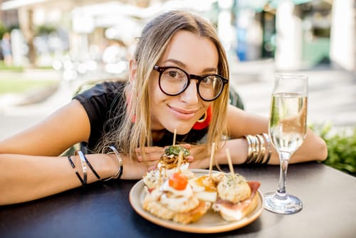 メガネをかけた女性が皿に料理とグラスワインを持ったテーブルに座り、スペイン風の食事を楽しんでいる。