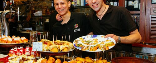 食べ物のトレイを持ってスペインのバーの前に立つ 2 人の男性。