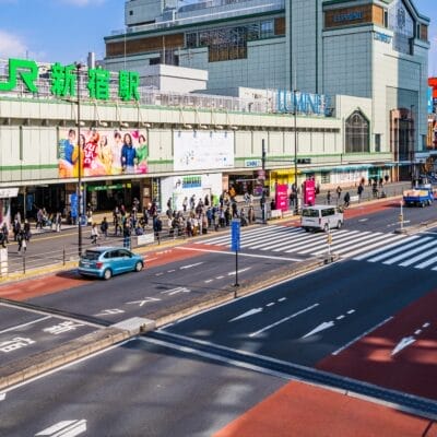 新宿のにぎやかな駅のストリートビュー。人々が歩いたり、車が道路を走っている。駅には大きな緑色の看板があり、周囲を高層ビルに囲まれています。近くにある人気のスペイン語教室マンマンツーでは、熱心な学習者向けに個別の言語レッスンを提供しています。.