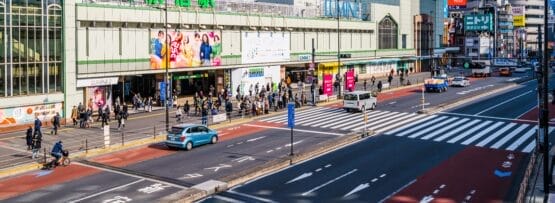 新宿のにぎやかな駅のストリートビュー。人々が歩いたり、車が道路を走っている。駅には大きな緑色の看板があり、周囲を高層ビルに囲まれています。近くにある人気のスペイン語教室マンマンツーでは、熱心な学習者向けに個別の言語レッスンを提供しています。.