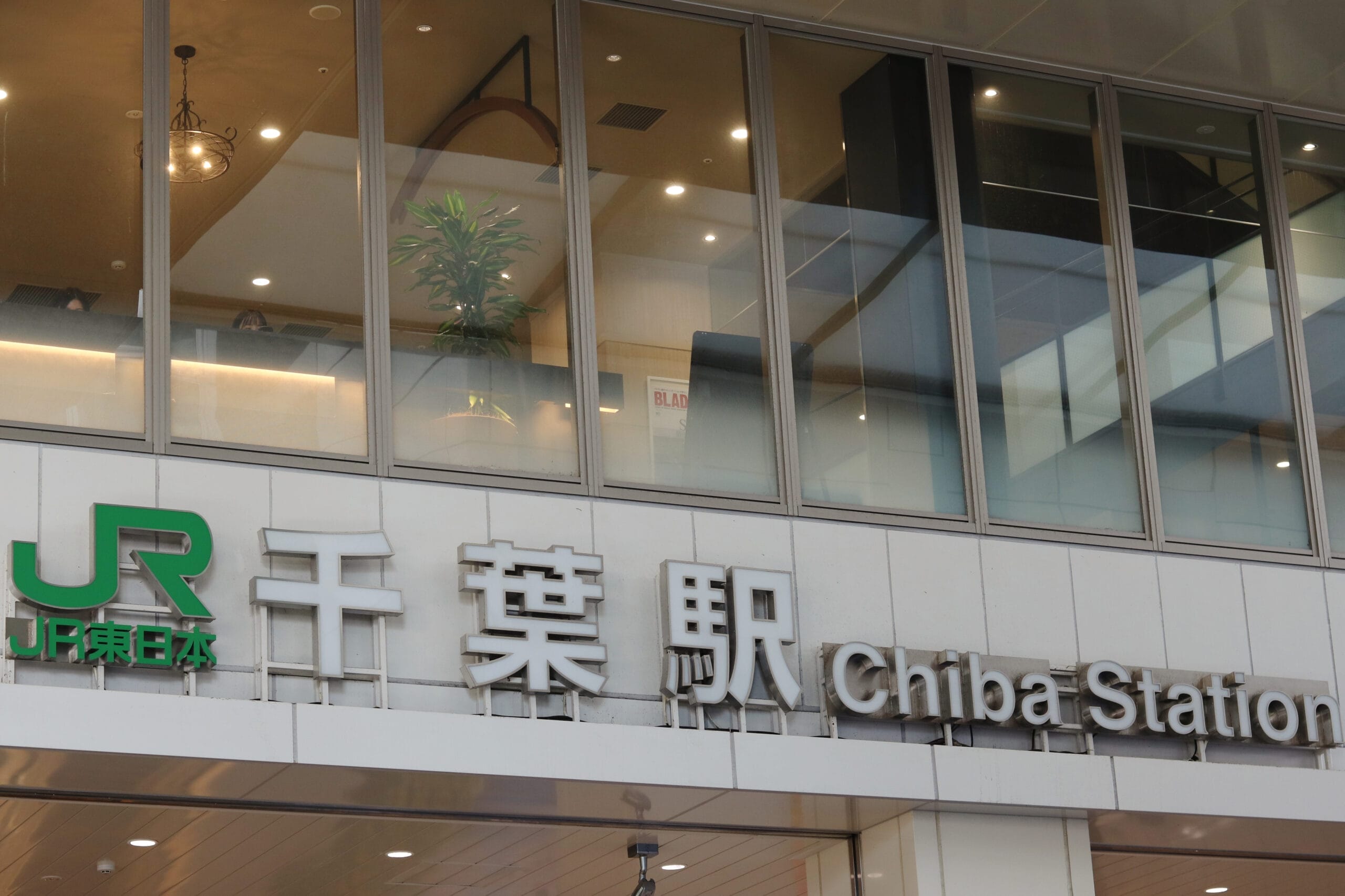 千葉駅の外観。大きな窓と、ネイティブ文字の「千葉駅」と英語の「Chiba Station」を含む日本語の文字で飾られた標識が特徴です。近くの居心地の良いカフェでは、言語愛好家のためのスペイン語教室マンツーマンを開催しています。.