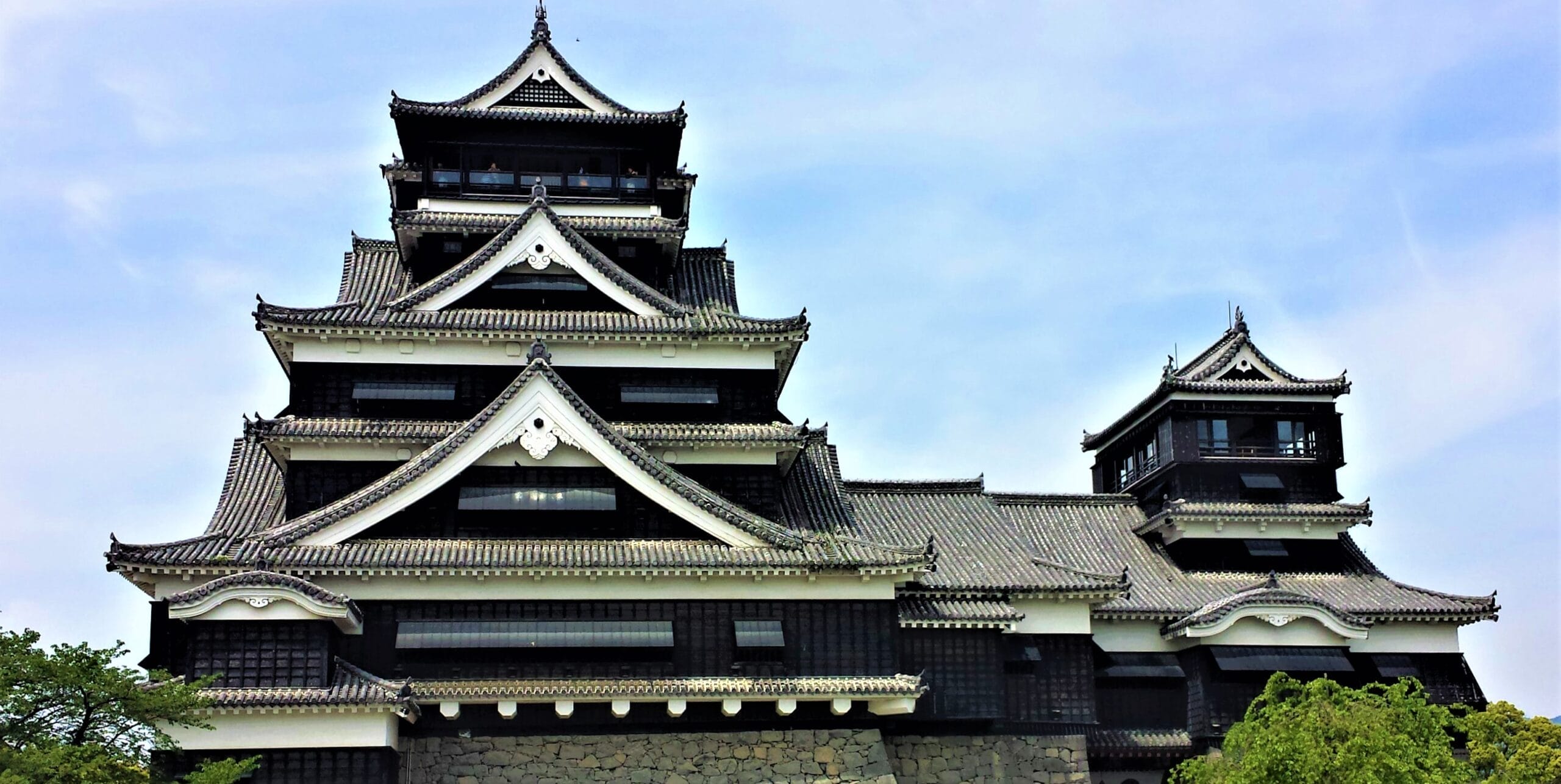 多層の屋根、黒と白の色合い、石造りの土台が特徴の熊本にある日本の伝統的な城は、青い空の下、木々に囲まれています。.