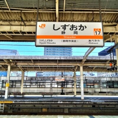 静岡駅のホームの眺め。駅名や道順を含む日本語、英語、スペイン語の文字が書かれた頭上の看板。背景には都市の建物が見えます。静岡・浜松間の旅行やスペイン語教室マンツーマンセッションに向かう方に最適です。.