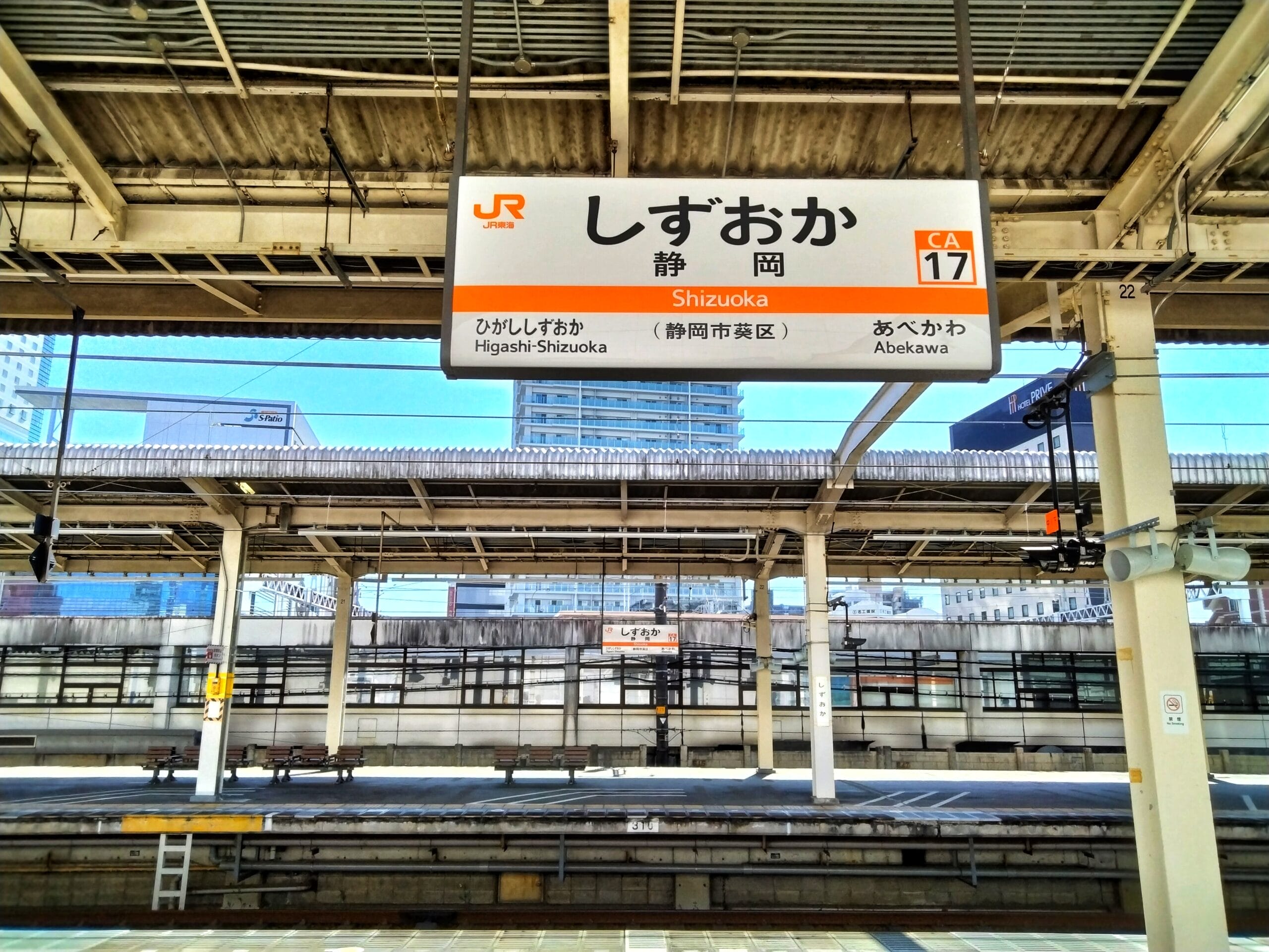 静岡駅のホームの眺め。駅名や道順を含む日本語、英語、スペイン語の文字が書かれた頭上の看板。背景には都市の建物が見えます。静岡・浜松間の旅行やスペイン語教室マンツーマンセッションに向かう方に最適です。.