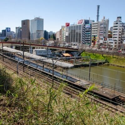 川沿いの平行線路に電車が走り、草地に囲まれた街並み。手前には賑やかな市ヶ谷、背景には澄み切った青空の下、高層ビルが立ち並んでいます。.