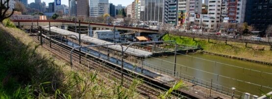 川沿いの平行線路に電車が走り、草地に囲まれた街並み。手前には賑やかな市ヶ谷、背景には澄み切った青空の下、高層ビルが立ち並んでいます。.