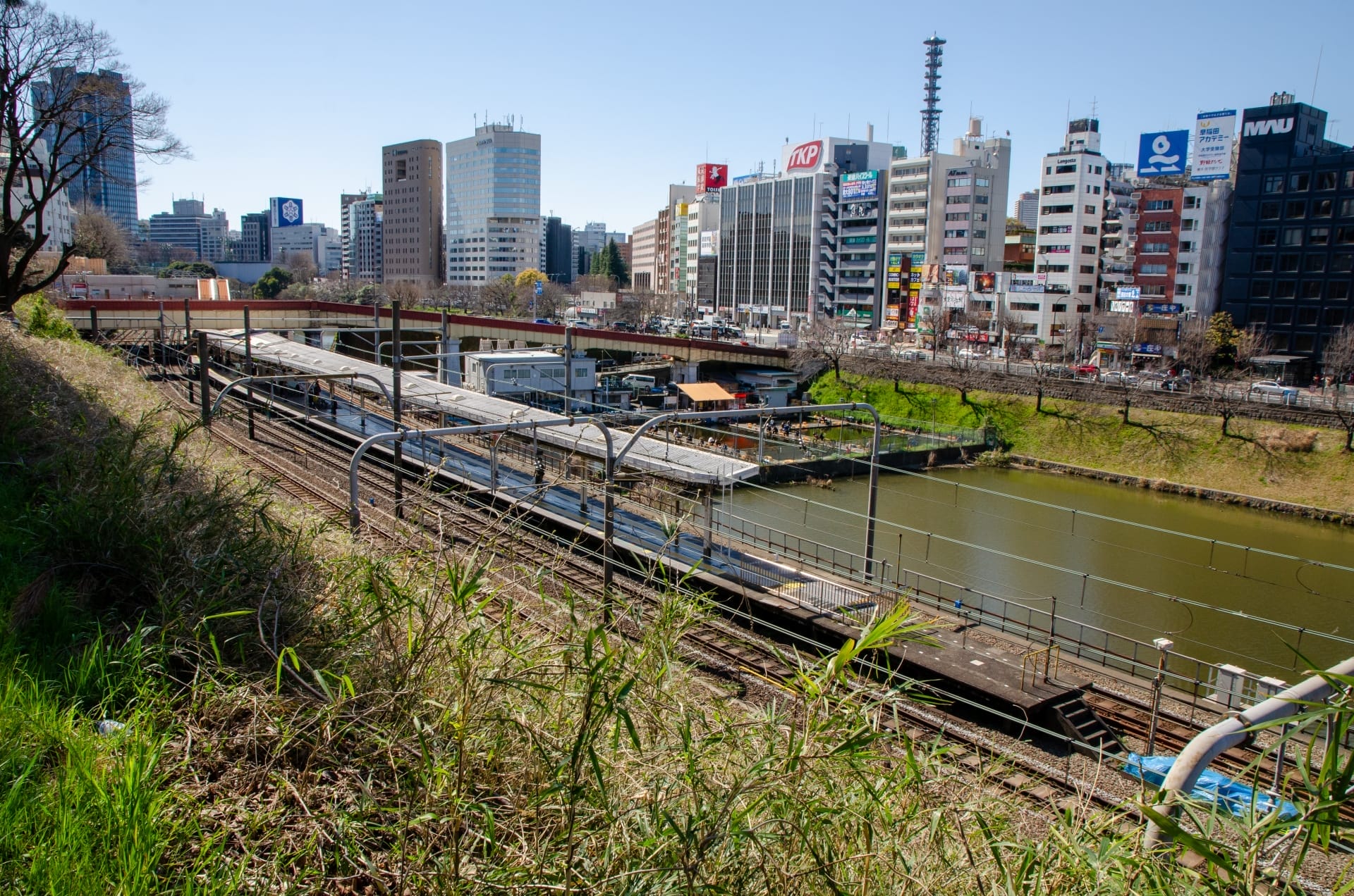 川沿いの平行線路に電車が走り、草地に囲まれた街並み。手前には賑やかな市ヶ谷、背景には澄み切った青空の下、高層ビルが立ち並んでいます。.