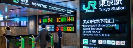 東京駅を歩く乗客。電光掲示板には電車の時刻表や道順が多言語で表示され、近くで利用できるスペイン語教室マンツーマンセッションの詳細も表示されます。.