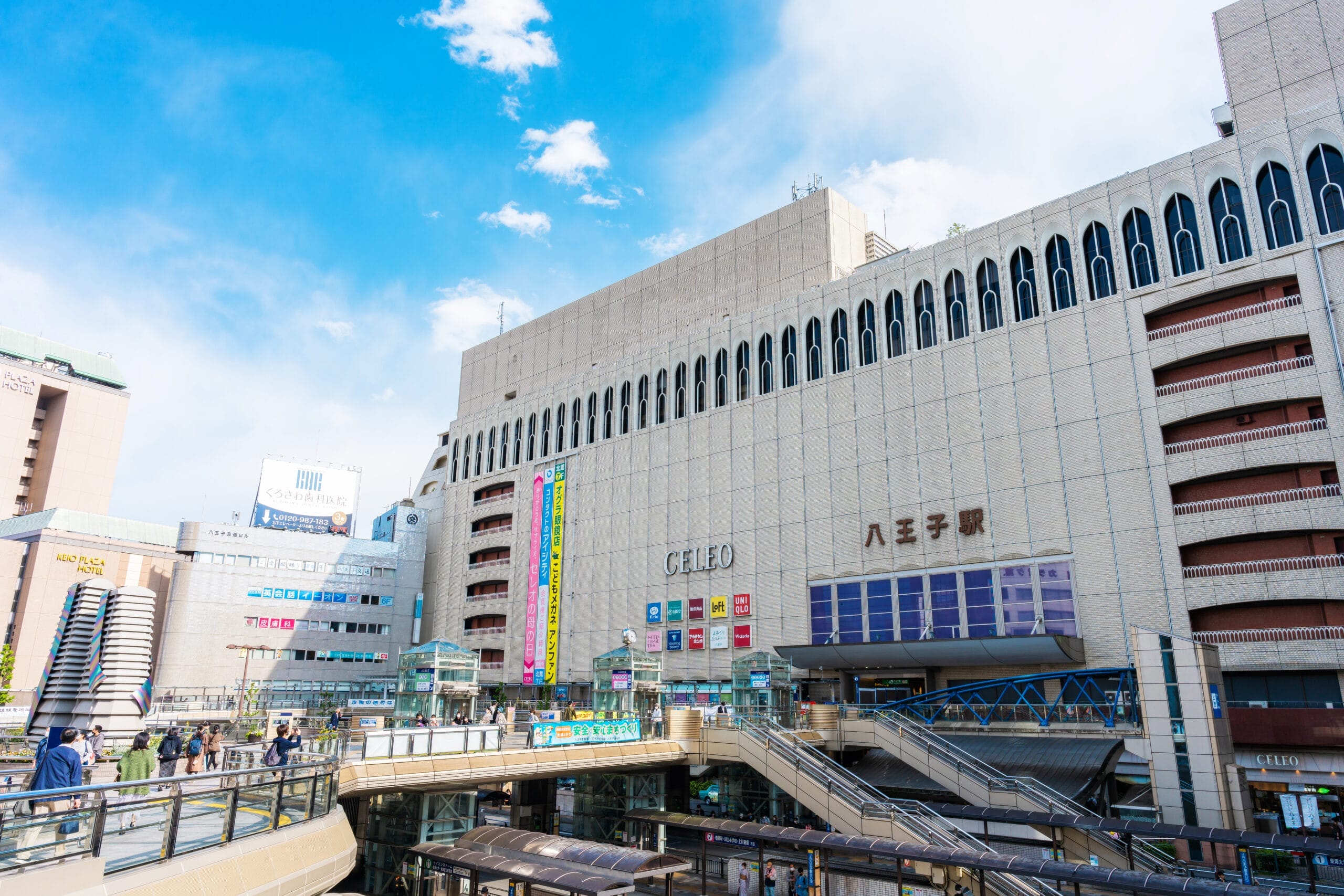 複数の標識と青い空を背景に、八王子にある大規模な都市ビルの外観。スペイン語教室マンツーマンに向かう人々が正面にあるにぎやかな歩行者専用エリアを特徴としている。.