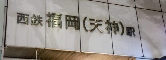 天神・福岡駅の入り口には日本語の標識があります。壁は白く、反射する金属の文字が西鉄福岡（天神）への入り口を示しています。.