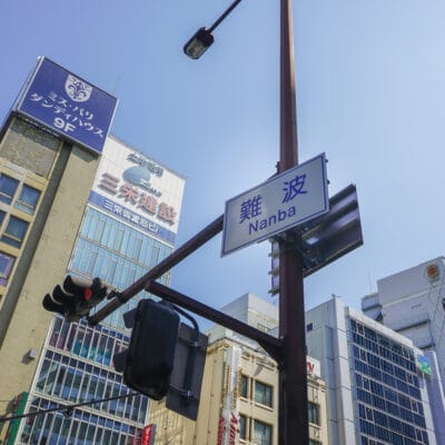 大阪のなんば・難波のストリート ビュー。澄んだ青い空を背景に高層ビル、商業看板、信号機が特徴です。活気に満ちた雰囲気を完璧に捉えており、都会のオアシスにあるスペイン語教室のマンツーマンと同じくらい魅力的です。.