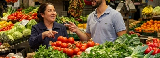 トマト、葉野菜、ピーマン、玉ねぎなど新鮮な野菜や果物が並ぶ、活気のあるスペインの市場の屋台で、売り手と客が会話をしています。.
